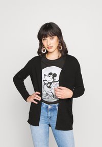Svart cardigan med lange ermer og åpen front, båret over en grafisk t-skjorte. Har lommer og et mykt, teksturert strikkemateriale.