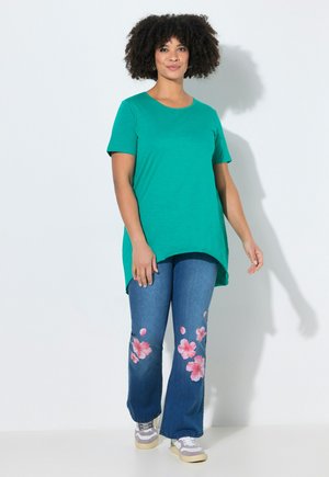 Frau steht und trägt ein grünes Kurzarmshirt sowie blaue Jeans mit pinkfarbenen Blumenmustern, kombiniert mit grauen und weißen Turnschuhen.