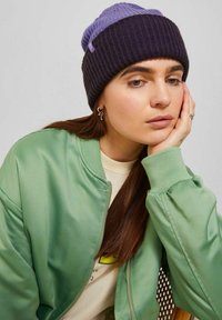 Beanie lavorato a maglia in blu scuro e viola chiaro, con un design a coste. Il modello indossa una giacca bomber in satin verde con una felpa beige sotto.