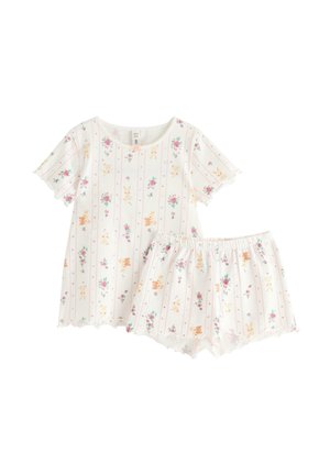 Ung pige står barfodet iført hvidt pyjamasæt med blomster- og kaninprint, korte ærmer og flæsekant.