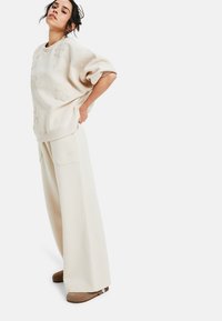 Vrouw poseert in een beige oversized sweatshirt met bloemembossing, wijde broek en bruine instappers tegen een witte achtergrond.
