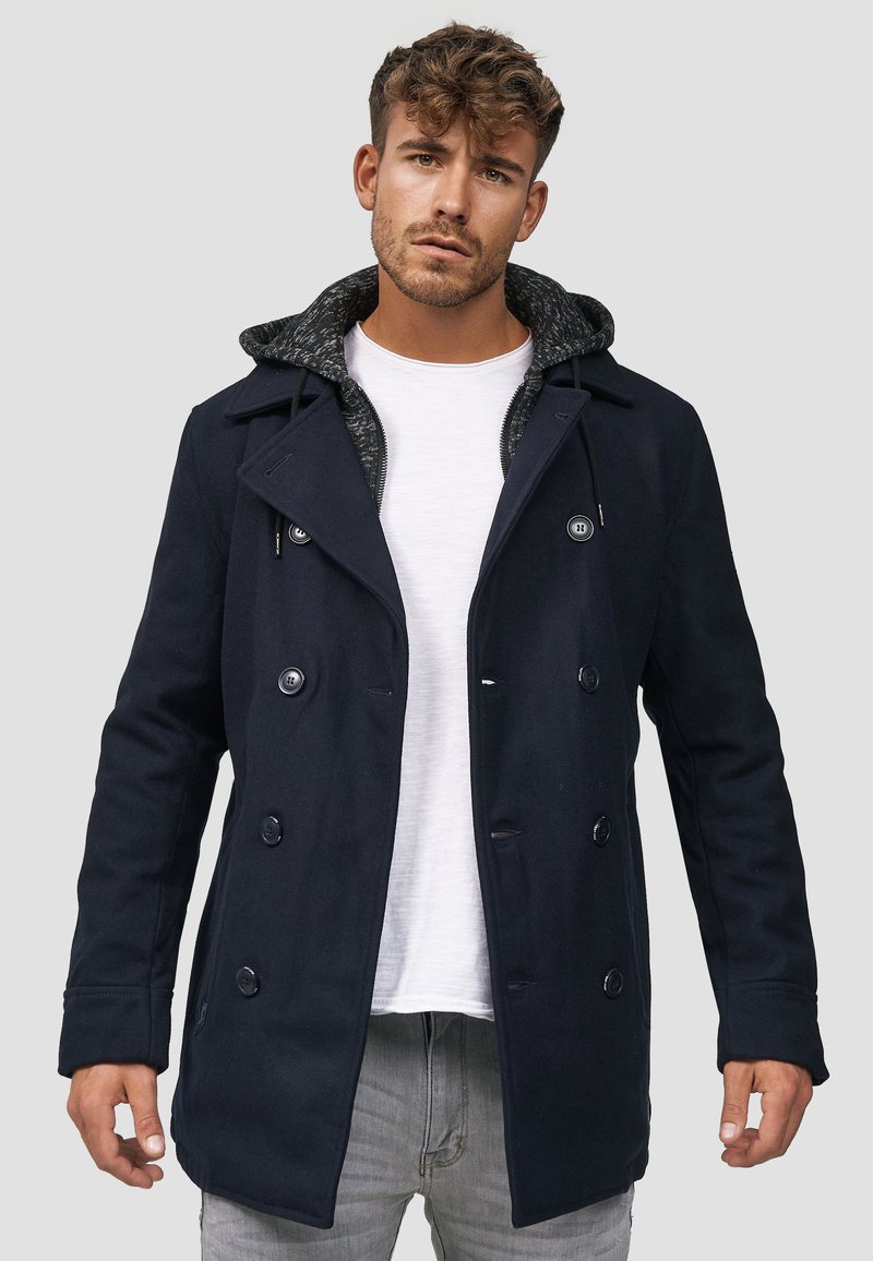 Marine peacoat med dobbeltknappede knapper og en grå mønstret hetteforing. Har en strukturert design og en midtlengde kutt.