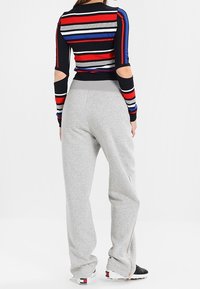 Langärmeliges Oberteil mit mehrfarbigen Streifen in Marineblau, Rot und Weiß; graue Jogginghose mit lockerem Schnitt und seitlichen Reißverschlüssen; sportliches Schuhwerk.
