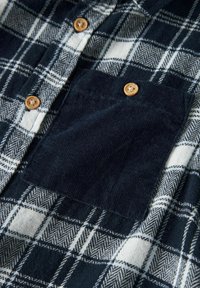 Rutete skjorte i marineblå og hvit, med en teksturert kordfløyelslomme, treknapper og et hundetannmønster. Myk stoff.