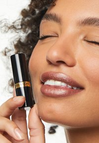 Rouge à lèvres Revlon dans un tube noir élégant avec une marque dorée. Le modèle a une peau douce et texturée et une couleur de lèvres naturelle avec une finition brillante.