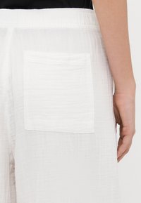 Bílé kalhoty z texturované tkaniny s elastickým pasem a jednou obdélníkovou náprsní kapsou na zadní straně. Hladký povrch, minimalistický design.