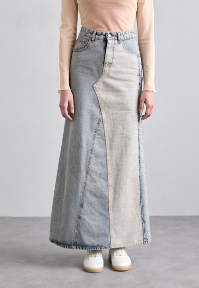 Jupe maxi en denim avec un design asymétrique unique, présentant deux nuances de bleu, un ourlet effiloché et un détail de couture à l'avant, associée à des baskets blanches.