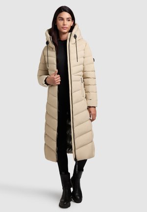 Beige langer Puffer-Mantel mit Kapuze, mit gestepptem Design, schwarzen Beschlägen und Reißverschluss vorne. Trägt über einem dunklen Outfit und schwarzen Stiefeln.