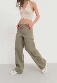 Béžový háčkovaný crop top s výstřihem do V, doplněný širokými olivově zelenými kalhotami typu cargo s bočními kapsami a předním zapínáním na knoflíky.