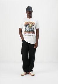 Hvid t-shirt med farverig "ADIDAS ORIGINALS" grafik, der viser en figur med hjelm, sammen med sorte brede bukser og hvide sneakers med røde striber.