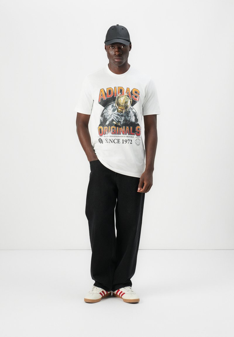 Camiseta blanca con un gráfico colorido de "ADIDAS ORIGINALS" que presenta una figura con casco, combinada con pantalones anchos negros y zapatillas blancas con rayas rojas.