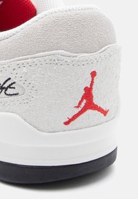 Λευκό αθλητικό παπούτσι με υφή από γκρι σουέτ στο τακούνι, κόκκινο λογότυπο Jumpman, μαύρη σόλα και λευκή μεσαία σόλα. Διαθέτει ραμμένα στοιχεία και επενδυμένο κολάρο.