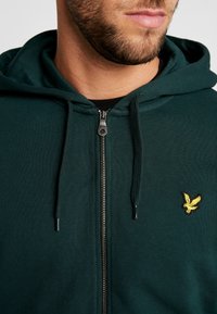 Tmavě zelená mikina na zip s klokankou, šňůrkami a malým žlutým ptáčkem na hrudi. Materiál je strukturovaný a měkký.