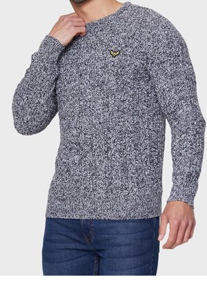Homme portant un pull tricoté noir et blanc avec un petit emblème jaune sur la poitrine, associé à un jean bleu, main près du cou.