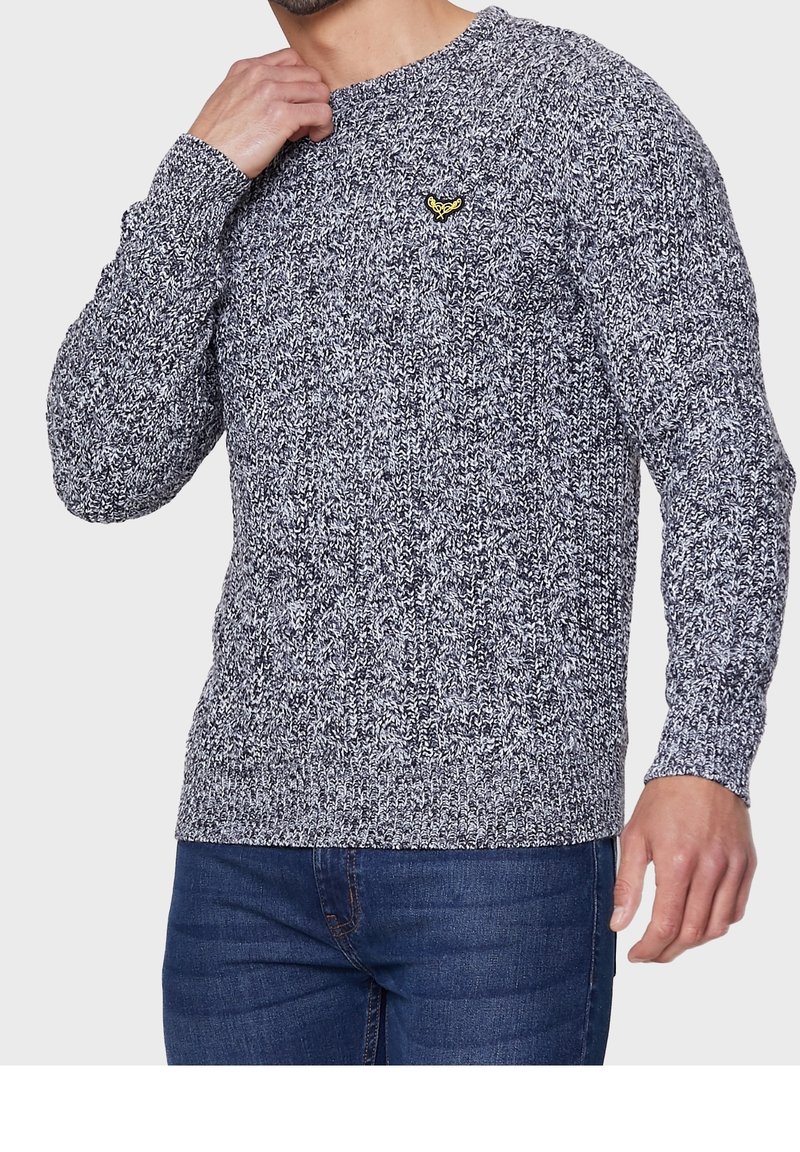 Homme portant un pull tricoté noir et blanc avec un petit emblème jaune sur la poitrine, associé à un jean bleu, main près du cou.