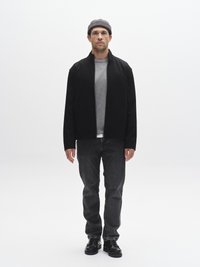 Juoda striukė ant pilkos marškinėlių, derinama su tamsiomis džinsinėmis kelnėmis. Pilka žieminė kepurė ir juodi batai užbaigia įvaizdį. Paprastas, struktūruotas dizainas ir neutralios spalvos.