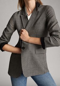 Harmaa houndstooth-takkia mustilla yksityiskohdilla, kahdella napilla ja muotoiltu istuvuus. Ominaisuuksiin kuuluvat lovetut kaulukset ja käärityt hihat.