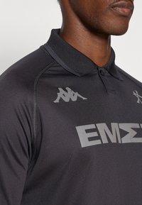 Svart pikétröja av texturerat tyg. Har en krage, grå logotyper och bokstäver samt subtila sömnaddetaljer.