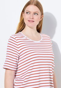 Bílý tričko s horizontálními červenými pruhy, kulatým výstřihem a krátkými rukávy. Měkká látka s uvolněným střihem. Jasné visací náušnice viditelné.
