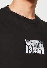 Primer plano de una persona que lleva una camiseta de cuello redondo negra con un logo blanco de Calvin Klein en el pecho.