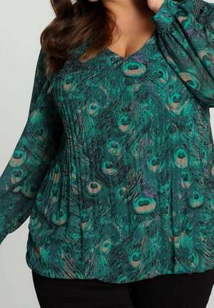 Kvinde iført grøn bluse med påfuglefjer-mønster og lange, gennemskinnelige ærmer, poserer med hånden nær ansigtet.