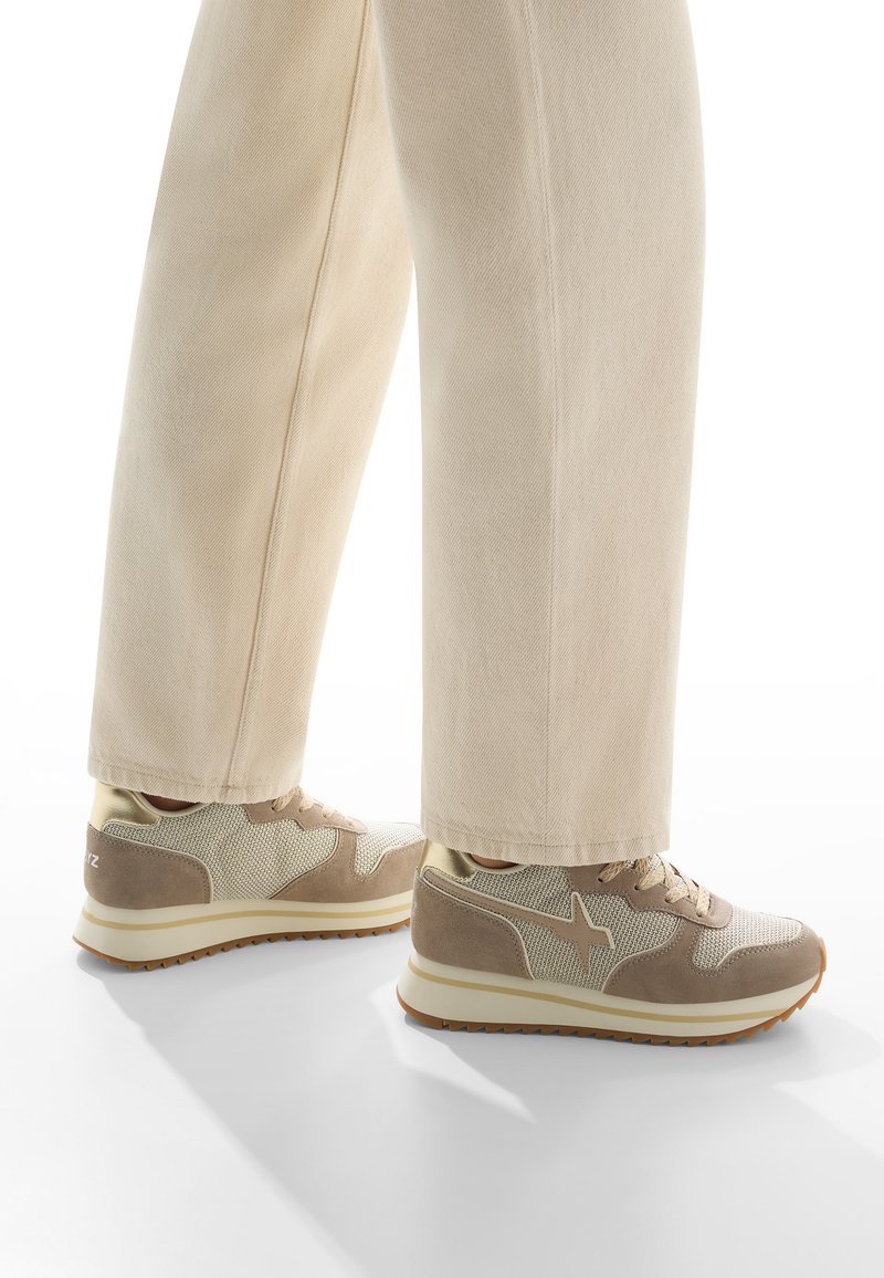 Sneakers beige in materiali mesh e suede, con suola bianca e dettagli marroni, abbinati a pantaloni larghi di colore chiaro.