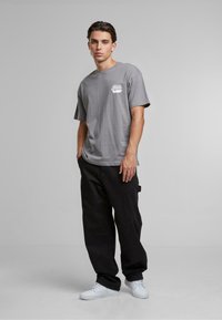 Light gray cotton t-shirt featuring a small white graphic, paired with loose black pants and white sneakers. Plain background.