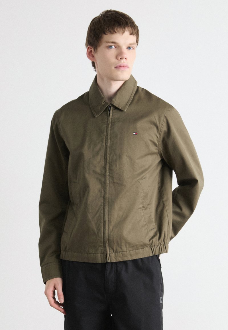 Oliven grønn zip-up jakke med krage, elastisk midje, sidelommer og liten logo akcent på venstre bryst. Lett stoff.