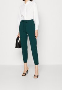 Kvinna i vit långärmad skjorta, teal höga midjebyxor med knyt, svarta högklackade sandaler, håller en liten svart handväska.