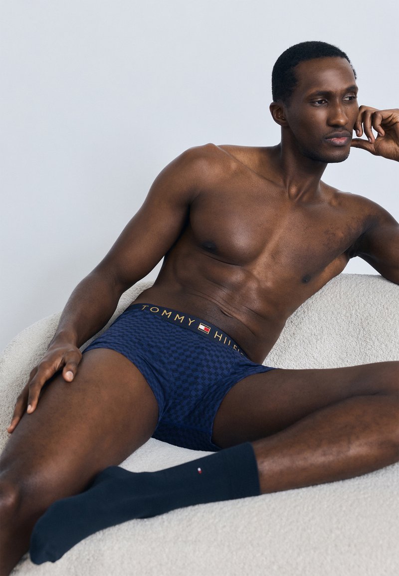 Sinise ruudulise mustriga bokserid kuldse logoga vöökohaga, mida kannavad tumesiniste ühevärviliste sokkidega, tekstuurse kreemja pinnaga taustal.