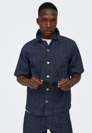 Homme portant une veste boutonnée à manches courtes bleu foncé et un pantalon assorti, tenant des lunettes de soleil noires des deux mains à hauteur de la taille.