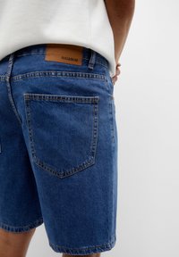 Pantalones cortos de mezclilla en azul oscuro con corte recto. Presenta dos bolsillos traseros y una etiqueta de marca de cuero marrón en la cinturilla.
