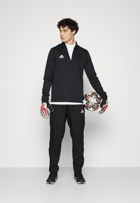 Top sportiv negru cu fermoar, guler alb, pantaloni negri, mănuși gri și o minge de fotbal cu un model, evidențiind o estetică sportivă modernă.