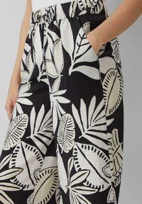 Personne portant un pantalon noir avec de grands motifs de feuilles blanches, la main dans la poche latérale, ceinture élastique visible.