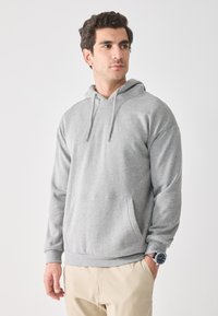 Hombre vestido con sudadera gris clara y pantalones beige, mirando hacia un lado, con una mano en el bolsillo y usando un reloj de pulsera.