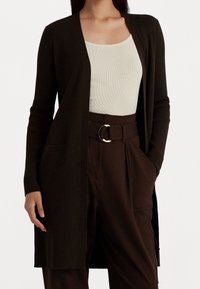 Cardigan lungo aperto in maglia a coste di un caldo marrone scuro, dotato di due tasche frontali e maniche lunghe, abbinato a una canotta beige chiaro e pantaloni marroni.