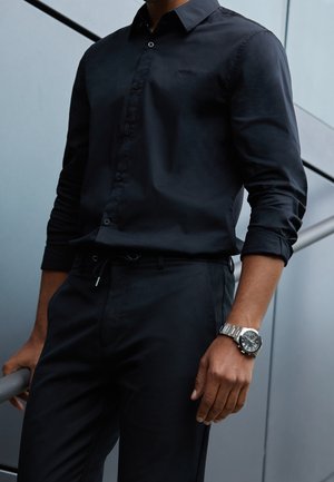 Homme portant une chemise noire boutonnée et un pantalon, une montre-bracelet argentée, debout près d'une rambarde en métal avec un mur gris en arrière-plan.