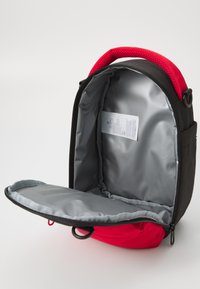 Svart og rød ryggsekk med et mesh-håndtak. Innvendig har den en grå foring og en flat lomme. Uoppslått viser den et romslig hovedrom.