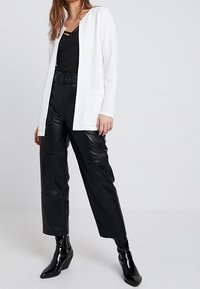 Cardigan bianco a maniche lunghe con apertura frontale, abbinato a una maglietta nera, pantaloni in pelle nera con cintura e stivaletti neri lucidi alla caviglia.