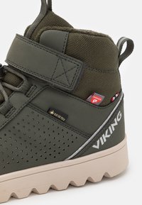 Botte de randonnée vert foncé avec une finition texturée, dotée d'une sangle Velcro, d'accents à pois et d'une semelle robuste de couleur claire. Étiquette waterproof visible.