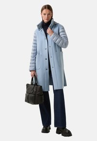 Frau in einem hellblauen Mantel mit gesteppten Ärmeln, dunklen Jeans, schwarzen klobigen Schuhen, die eine schwarze gepolsterte Handtasche hält und vor einem weißen Hintergrund steht.