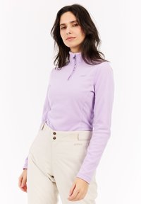 Donna che indossa un top in pile leggero viola chiaro con zip e pantaloni beige a vita alta su uno sfondo bianco uniforme.