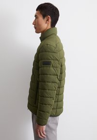 Olive green puffer jacket with quilted texture, high collar, side zipper pockets, and a logo patch on the shoulder.