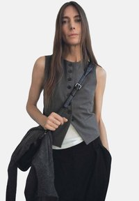 Gilet grigio senza maniche con chiusura a bottoni e due tasche, indossato su una maglietta bianca. Borsa a tracolla e sciarpa grigia tenute in una mano.