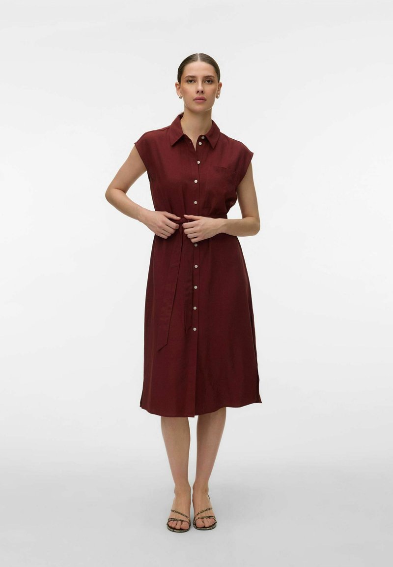 Femme portant une robe midi bordeaux sans manches à boutons, avec un col, une ceinture nouée et des sandales à bout ouvert, debout devant un fond uni.