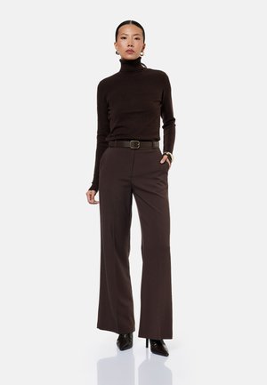 Nainen pukeutunut istuvaan tummanruskeaan poolokauluspuseroon, leveälahkeisiin ruskeisiin housuihin vyöllä, mustiin korkokenkiin ja kultaisiin rengaskorvakoruihin.