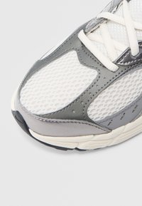 Γκρι και λευκό αθλητικό παπούτσι με πλεκτό επάνω μέρος, λείες δερμάτινες λεπτομέρειες και υφασμένη σόλα με ορατά μοτίβα πέλματος.