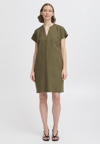 Vestido de lino verde oliva con escote en V, manga corta y corte holgado. Presenta un diseño de largo hasta la rodilla y una textura en la tela. Sandalias negras con tiras.