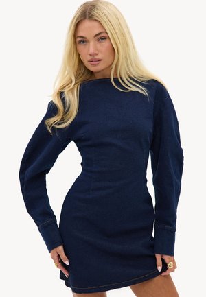 Blonde vrouw in een nauwsluitende donkerblauwe denim jurk met lange mouwen, poserend met handen bij de zoom tegen een witte achtergrond.