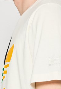 Triko Adidas v off-white bavlněném materiálu s grafikou obsahující žluté, zelené a černé prvky; na rukávu je vyšívané logo.